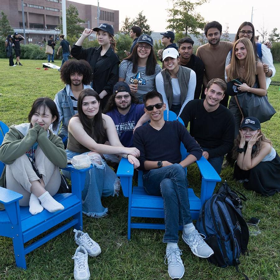 Group of students pose in blue chairs on campus center lawn.