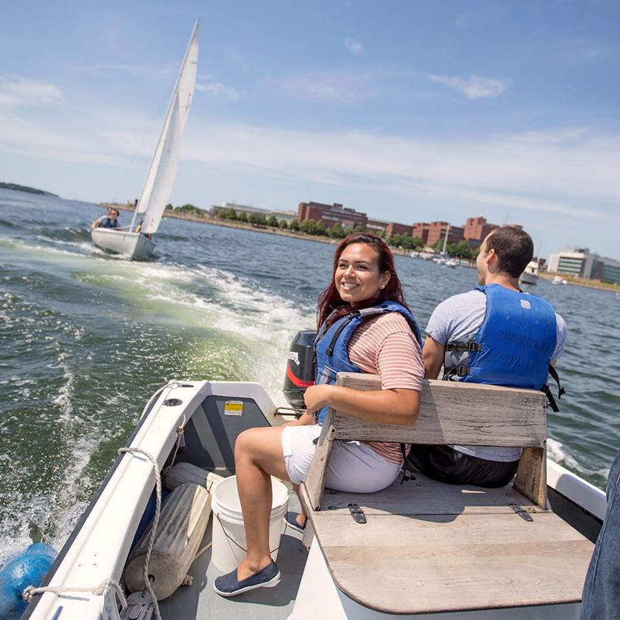 Two students on a motorboat with students on sailboat and campus in background.